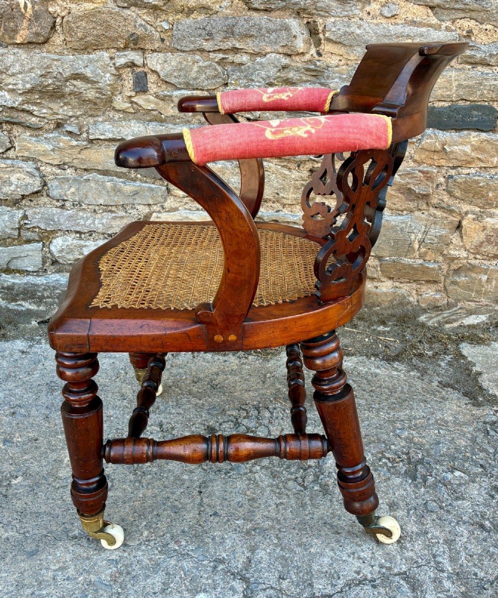 A Victorian mahogany desk chair featuring a curved top rail, pierced decorative splats, fabric-wrapped armrests, a woven cane seat, and turned legs with brass castors. View IV