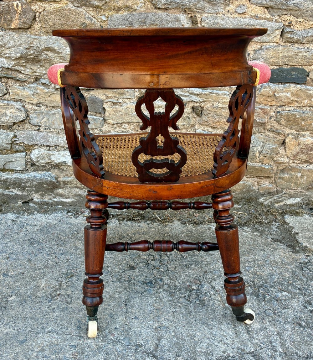 A Victorian mahogany desk chair featuring a curved top rail, pierced decorative splats, fabric-wrapped armrests, a woven cane seat, and turned legs with brass castors. View V