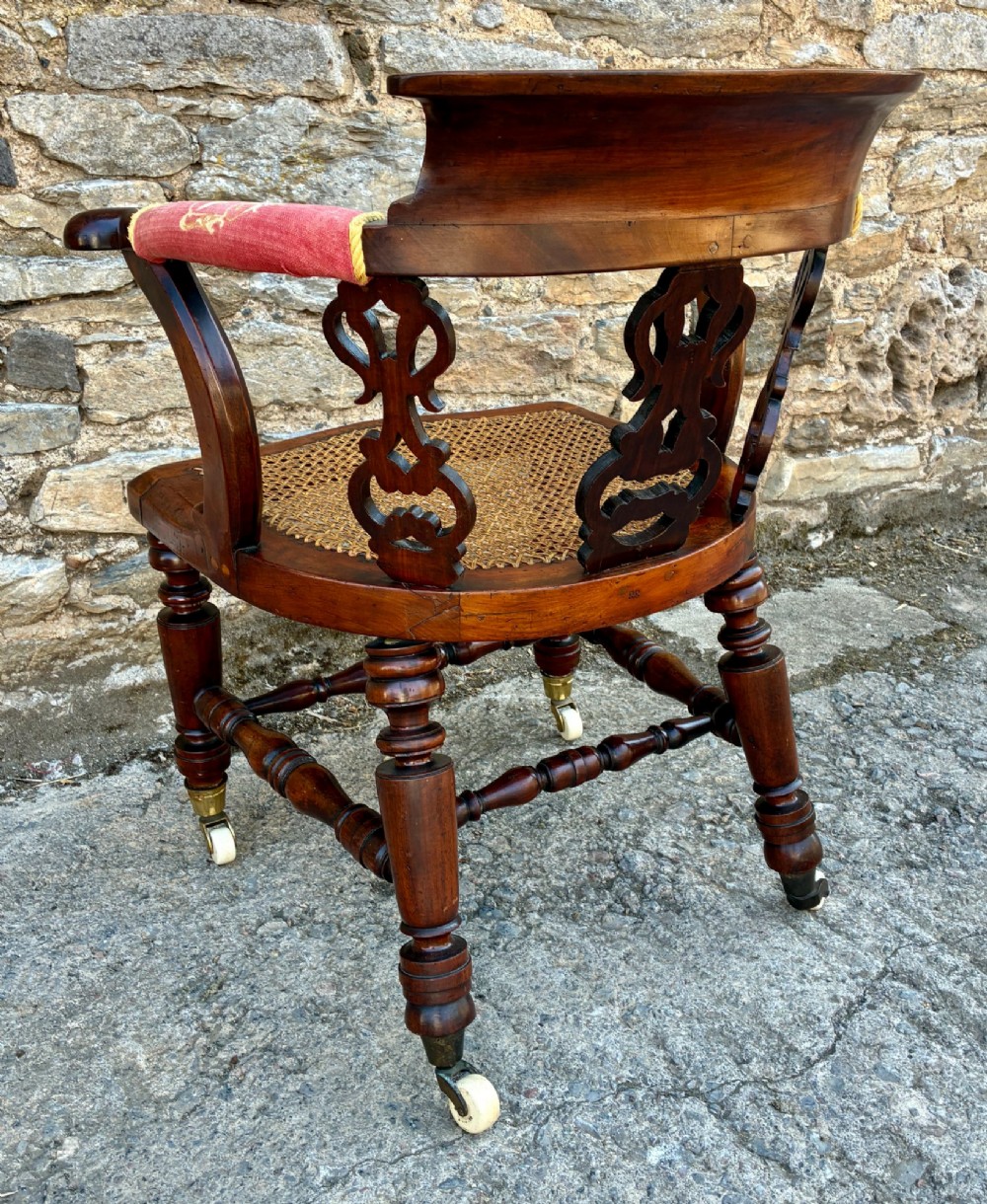 A Victorian mahogany desk chair featuring a curved top rail, pierced decorative splats, fabric-wrapped armrests, a woven cane seat, and turned legs with brass castors. View VI