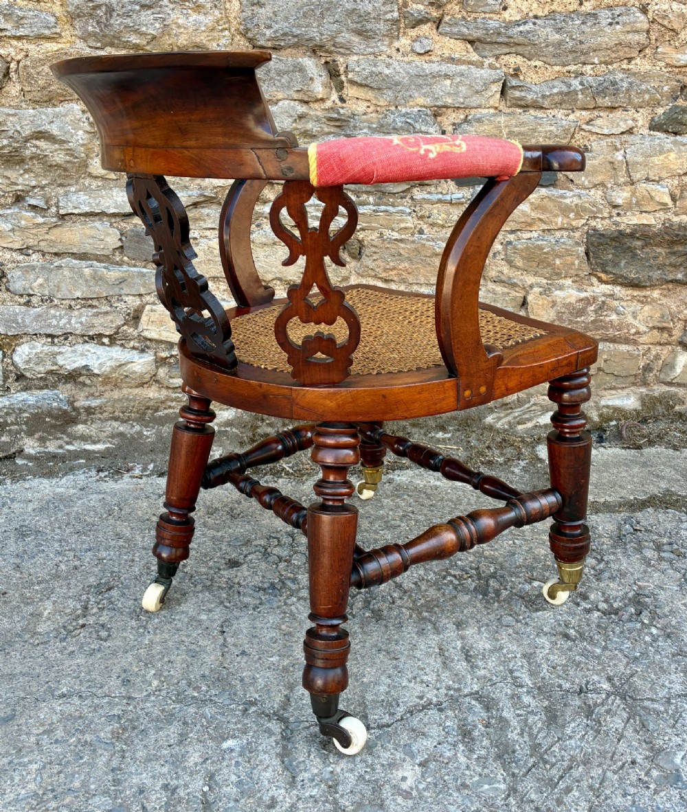 A Victorian mahogany desk chair featuring a curved top rail, pierced decorative splats, fabric-wrapped armrests, a woven cane seat, and turned legs with brass castors. View VIII