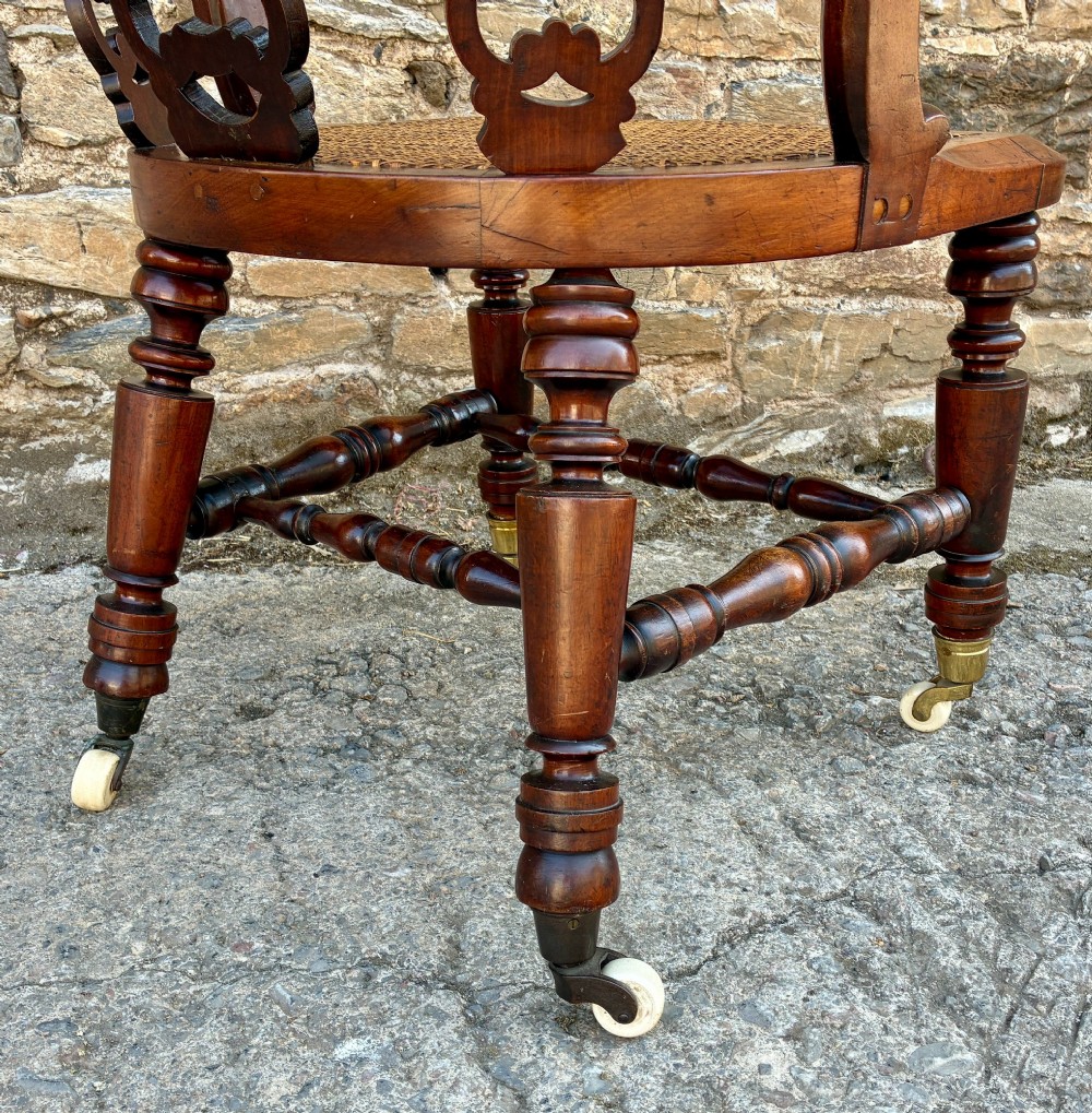 A Victorian mahogany desk chair featuring a curved top rail, pierced decorative splats, fabric-wrapped armrests, a woven cane seat, and turned legs with brass castors. View IX