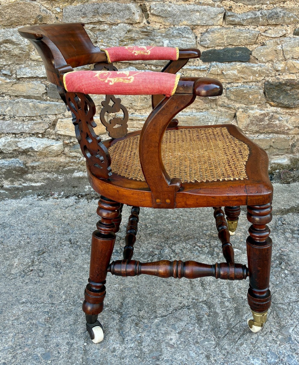 A Victorian mahogany desk chair featuring a curved top rail, pierced decorative splats, fabric-wrapped armrests, a woven cane seat, and turned legs with brass castors. View X