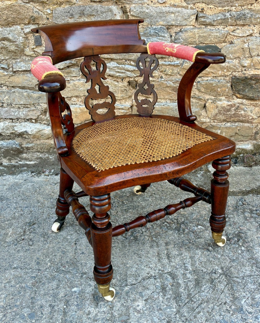 A Victorian mahogany desk chair featuring a curved top rail, pierced decorative splats, fabric-wrapped armrests, a woven cane seat, and turned legs with brass castors. View XI