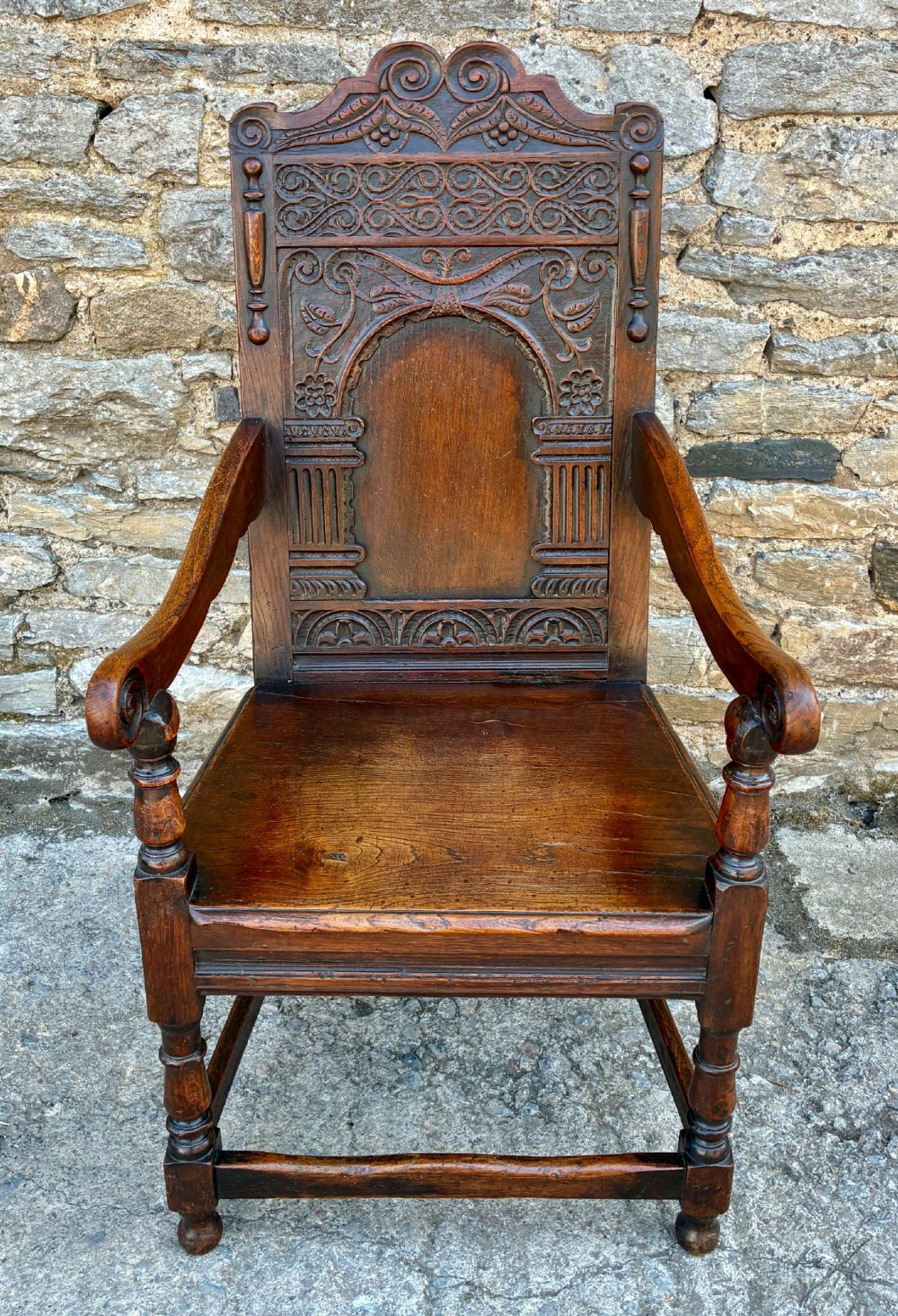 A dark carved oak wainscot armchair with a sculpted cresting rail, detailed panel back, turned front legs, and wooden stretchers.
