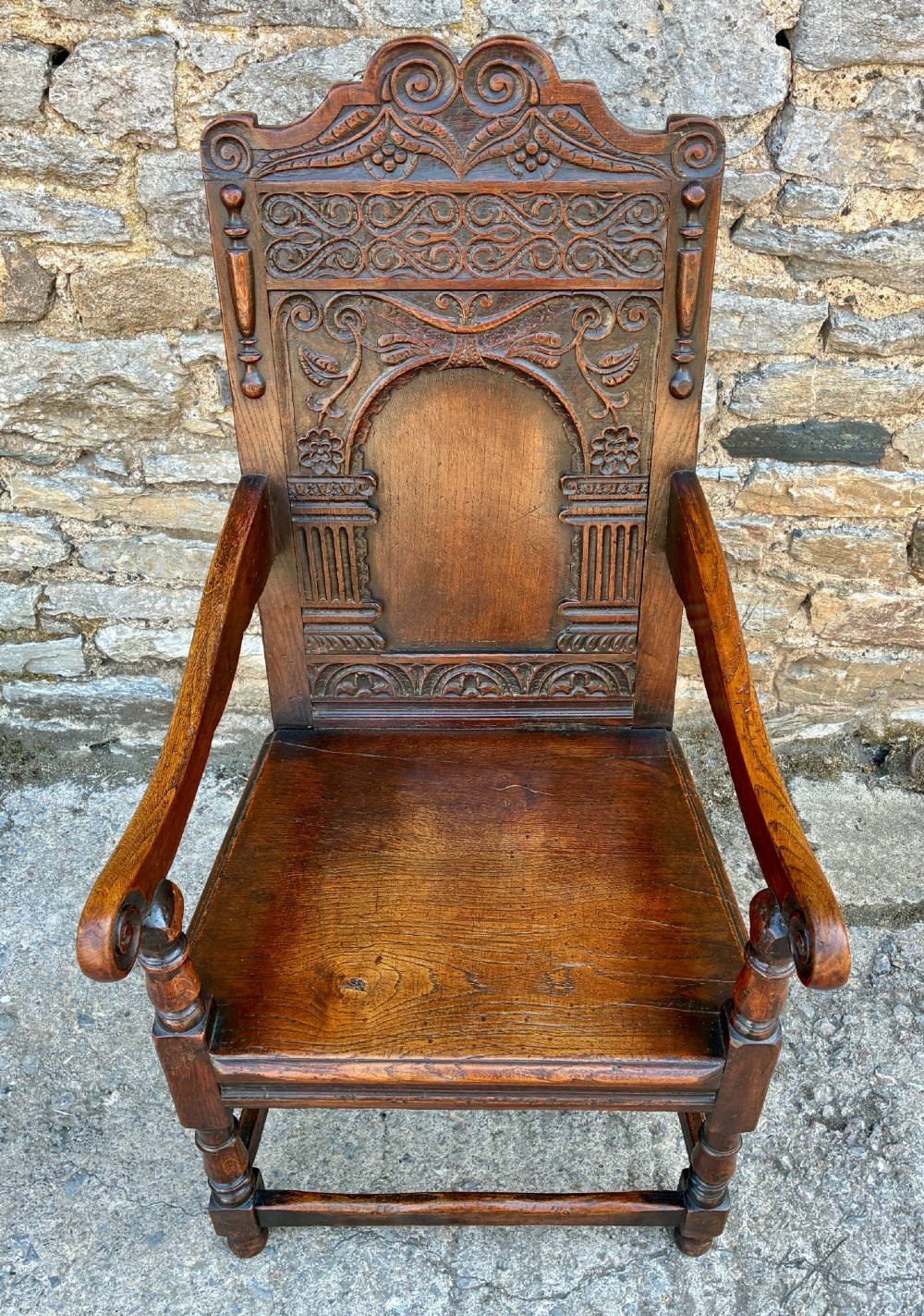 A dark carved oak wainscot armchair with a sculpted cresting rail, detailed panel back, turned front legs, and wooden stretchers. View II
