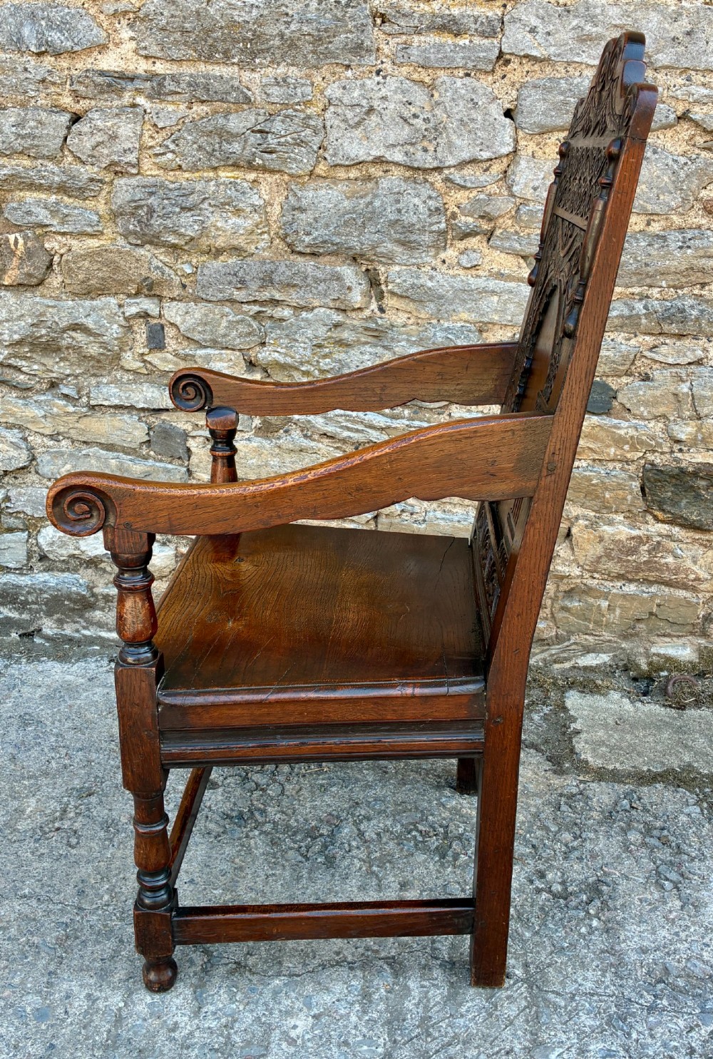 A dark carved oak wainscot armchair with a sculpted cresting rail, detailed panel back, turned front legs, and wooden stretchers. View V