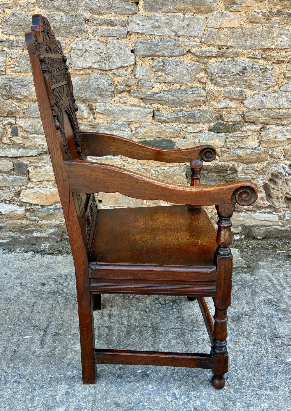 A dark carved oak wainscot armchair with a sculpted cresting rail, detailed panel back, turned front legs, and wooden stretchers. View IX