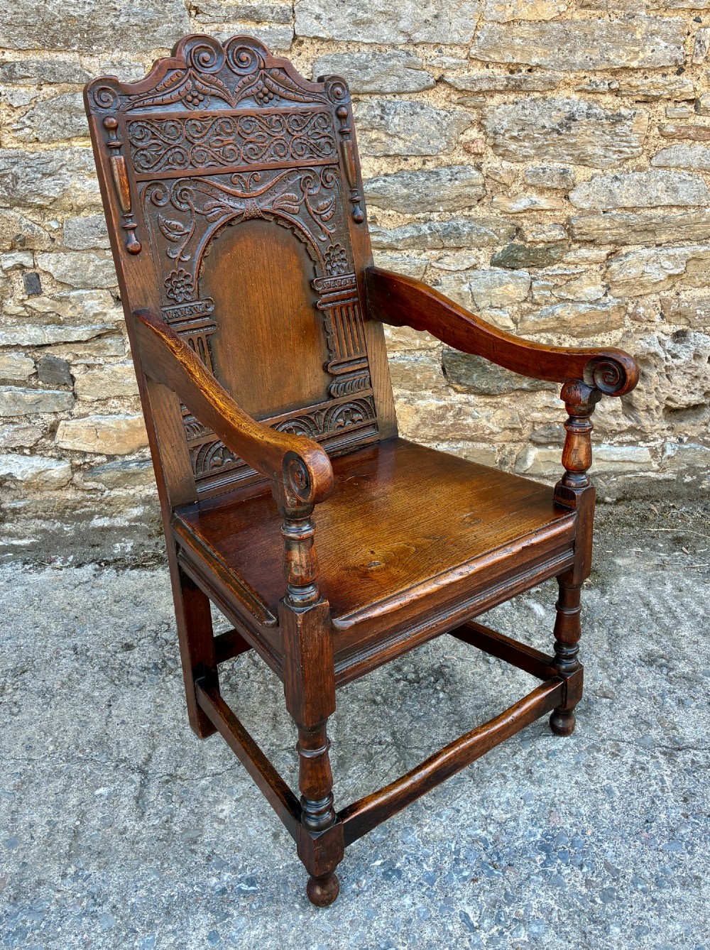 A dark carved oak wainscot armchair with a sculpted cresting rail, detailed panel back, turned front legs, and wooden stretchers. View X