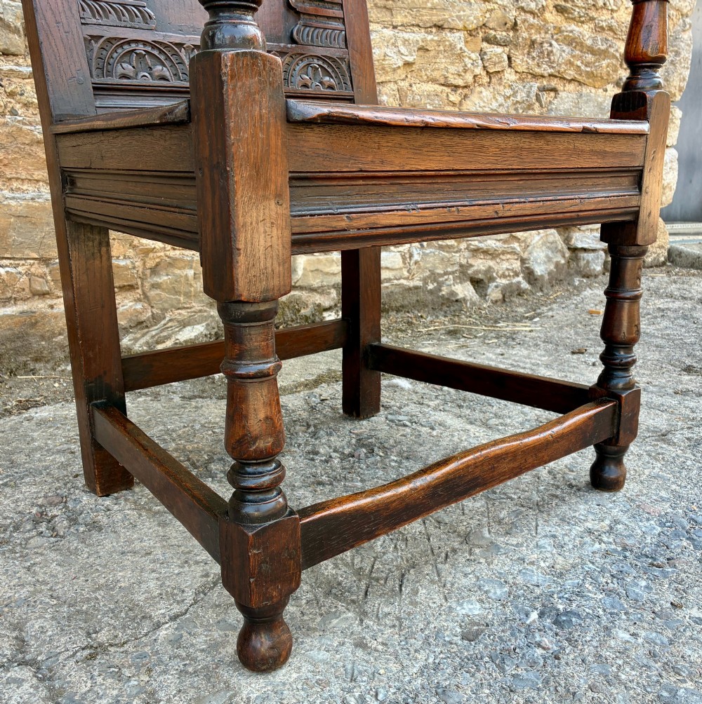 A dark carved oak wainscot armchair with a sculpted cresting rail, detailed panel back, turned front legs, and wooden stretchers. View XII