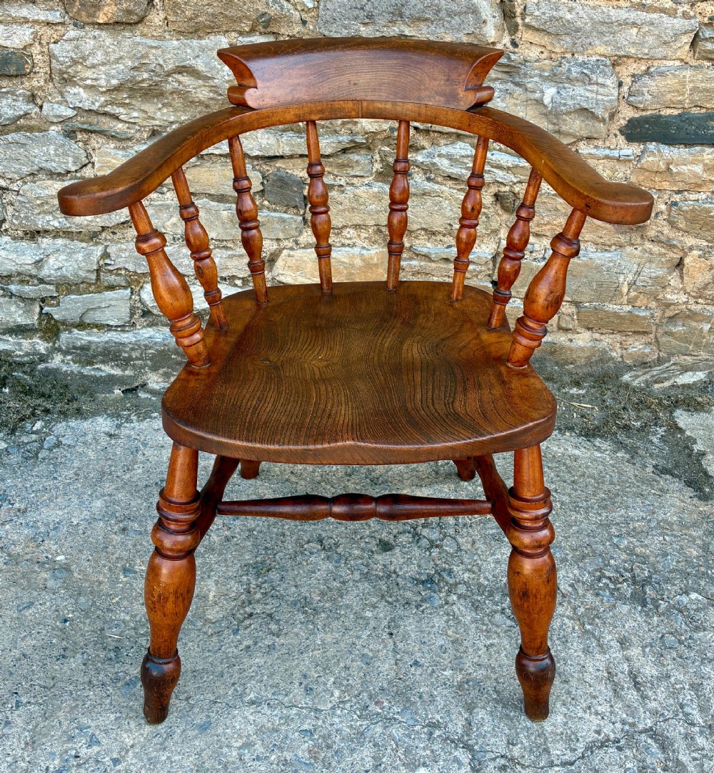 An antique wooden captains chair crafted from elm with turned spindles, a curved top rail, a contoured saddle seat, and turned legs.