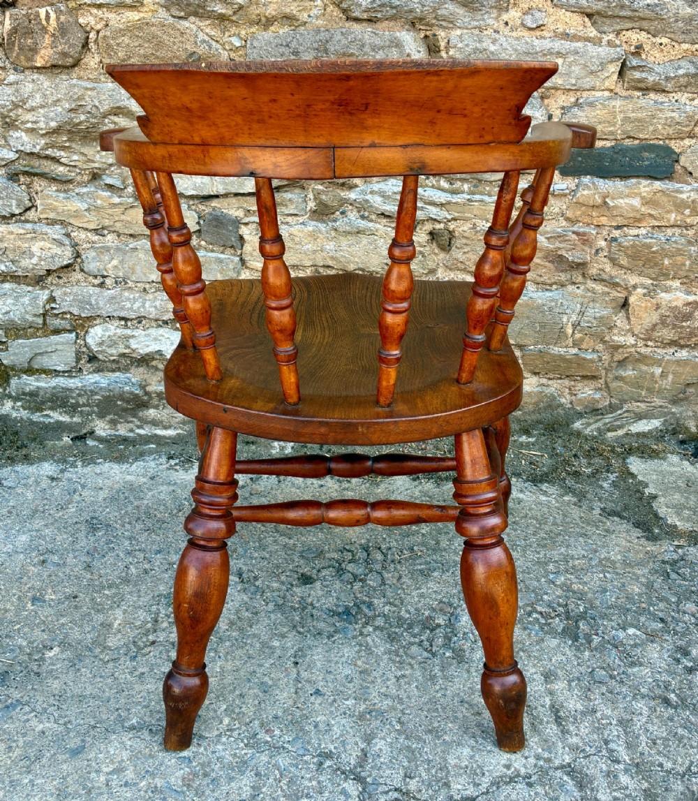 An antique wooden captains chair crafted from elm with turned spindles, a curved top rail, a contoured saddle seat, and turned legs. View V