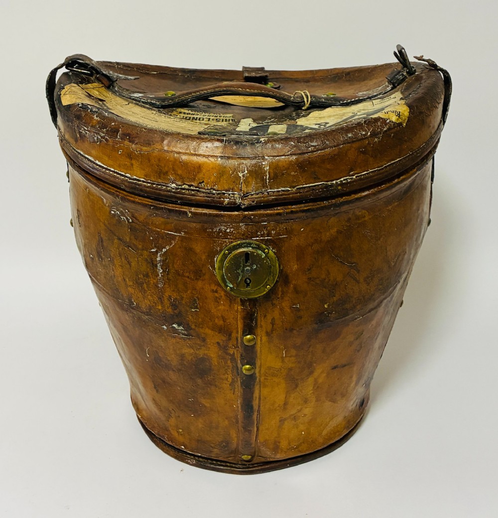 An antique tan leather double hat box featuring a shaped lid, top carrying handle, brass lock plate, and historical paper travel labels.