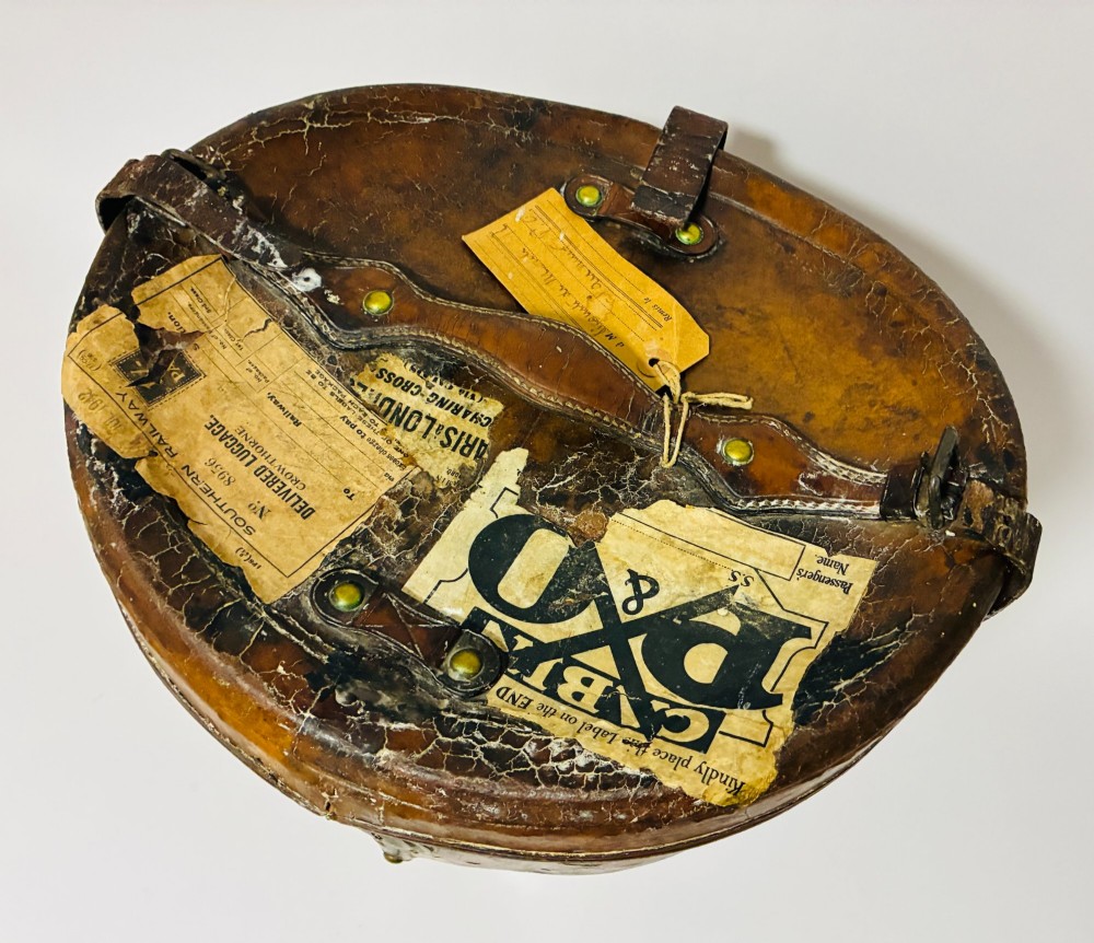 An antique tan leather double hat box featuring a shaped lid, top carrying handle, brass lock plate, and historical paper travel labels. View II