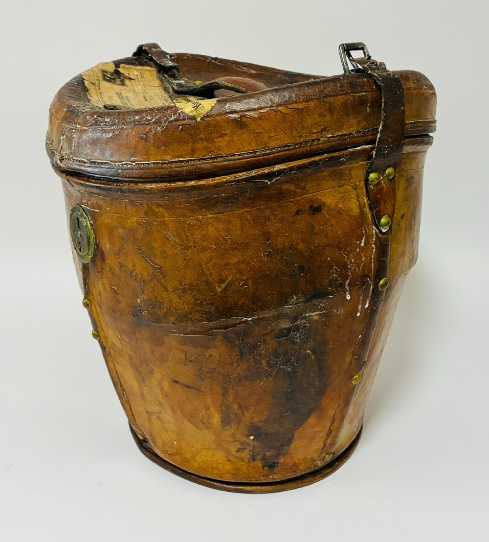 An antique tan leather double hat box featuring a shaped lid, top carrying handle, brass lock plate, and historical paper travel labels. View III