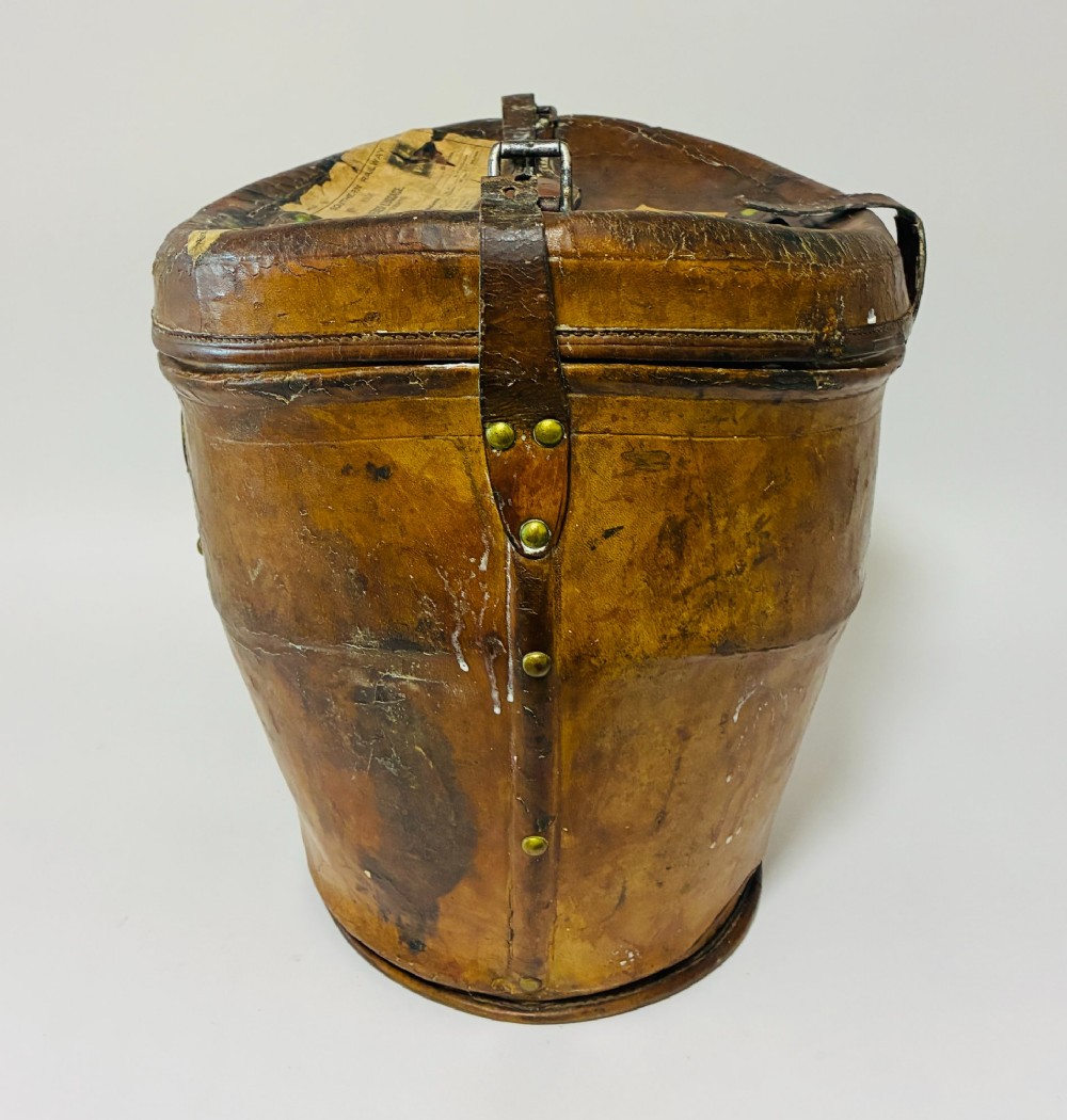An antique tan leather double hat box featuring a shaped lid, top carrying handle, brass lock plate, and historical paper travel labels. View IV