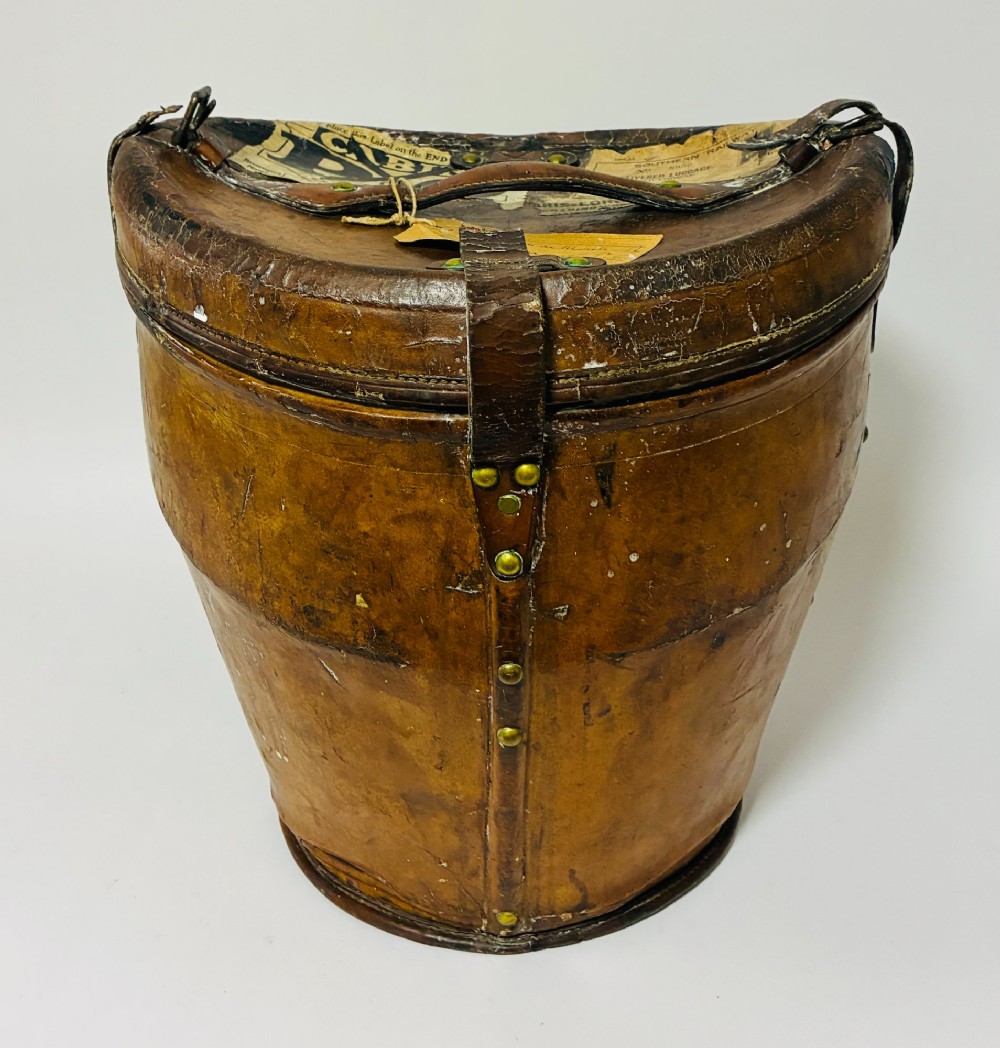 An antique tan leather double hat box featuring a shaped lid, top carrying handle, brass lock plate, and historical paper travel labels. View V