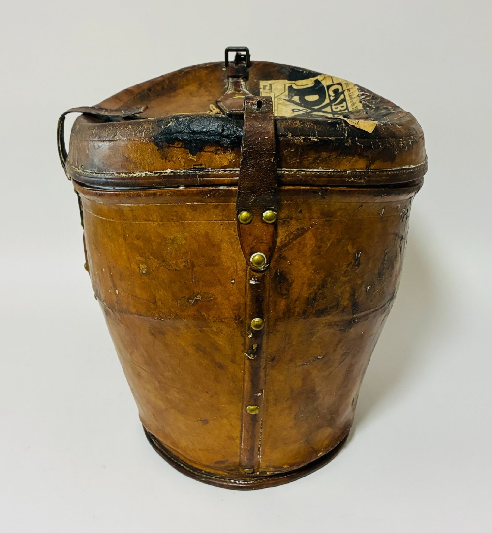 An antique tan leather double hat box featuring a shaped lid, top carrying handle, brass lock plate, and historical paper travel labels. View VI