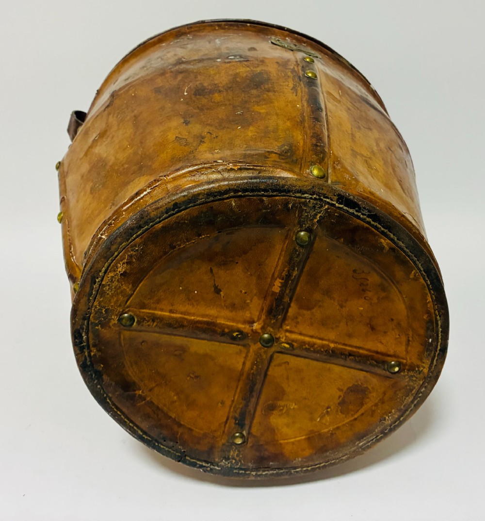 An antique tan leather double hat box featuring a shaped lid, top carrying handle, brass lock plate, and historical paper travel labels. View XV