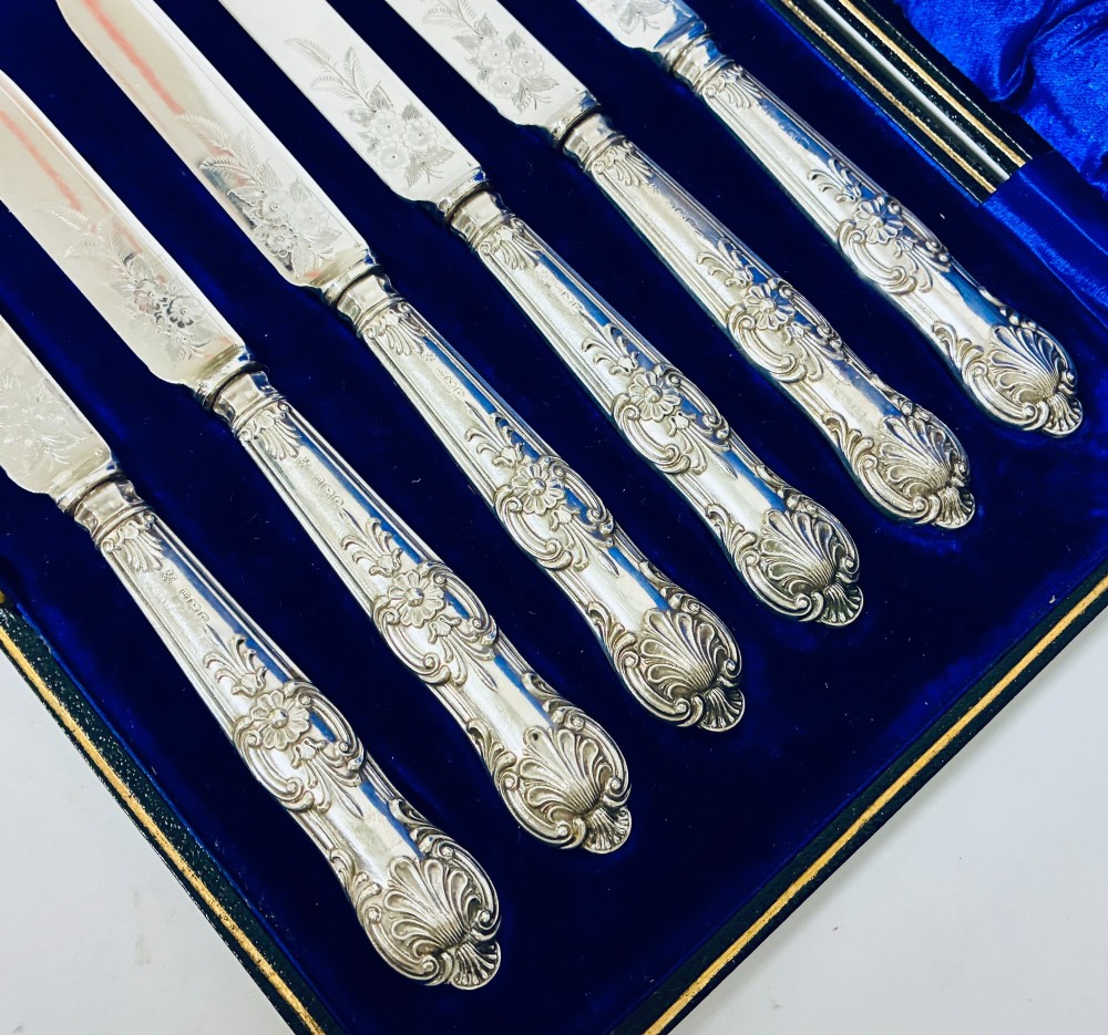 A cased set of six silver-handled fruit knives with floral engraved blades, displayed inside a blue velvet-lined presentation box. View III