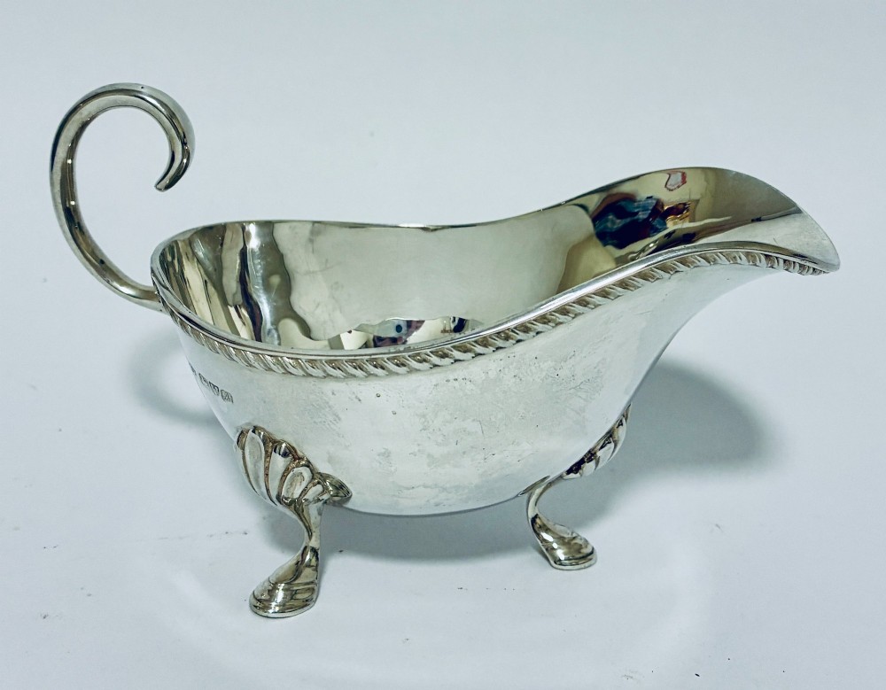 A polished sterling silver sauce boat featuring a flying scroll handle, gadrooned border, and three shell-capped pad feet.