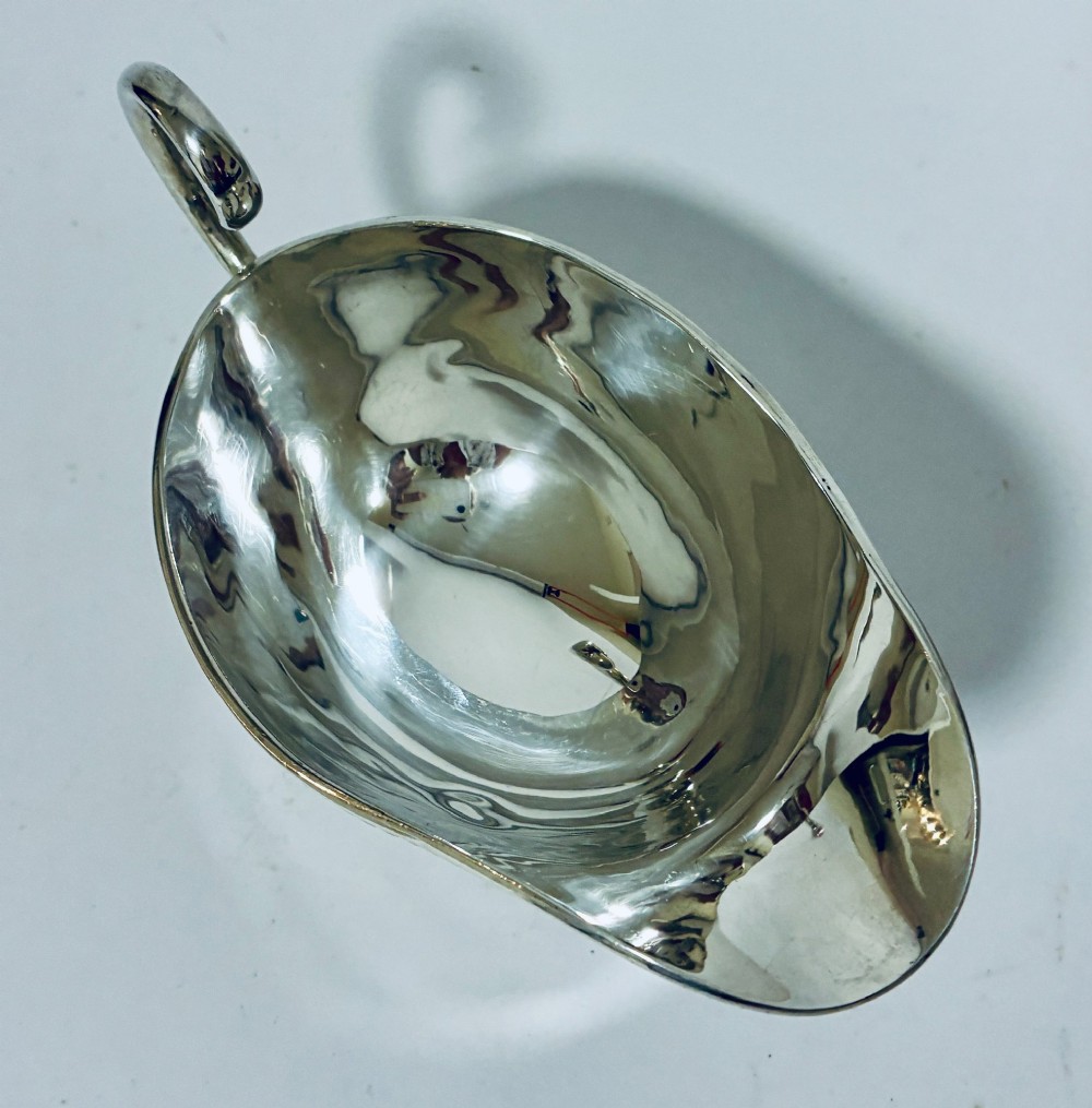 A polished sterling silver sauce boat featuring a flying scroll handle, gadrooned border, and three shell-capped pad feet. View VI