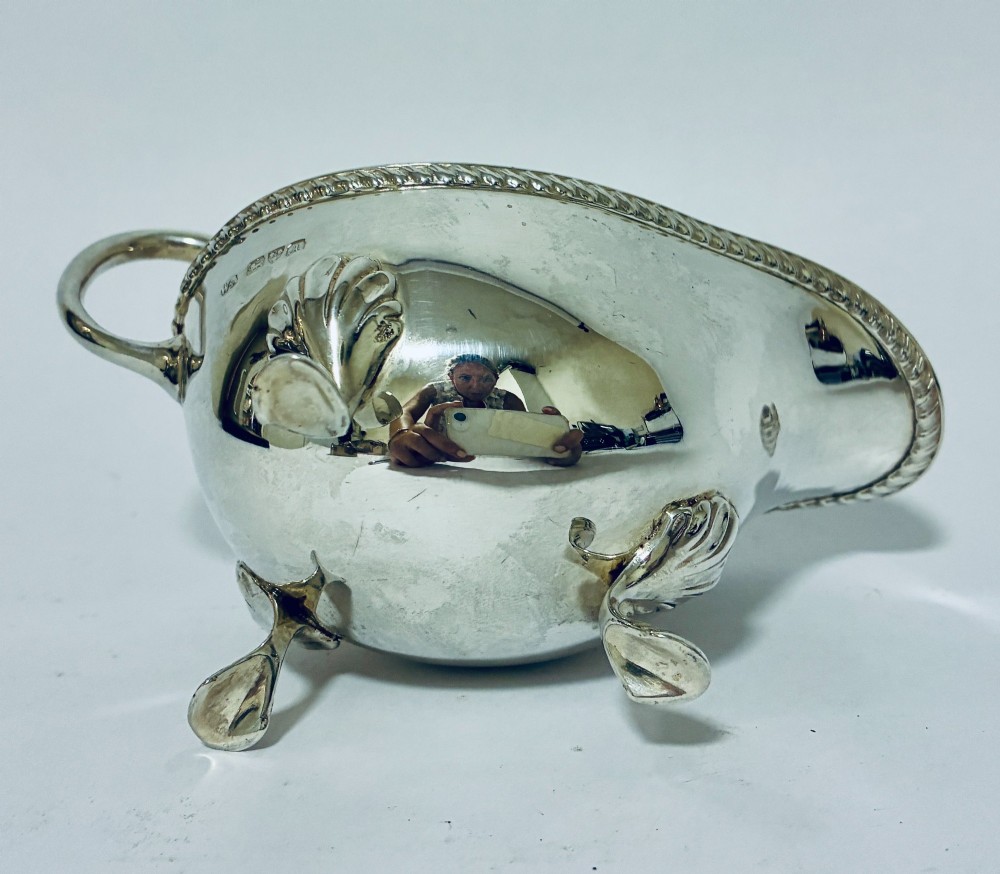 A polished sterling silver sauce boat featuring a flying scroll handle, gadrooned border, and three shell-capped pad feet. View VII