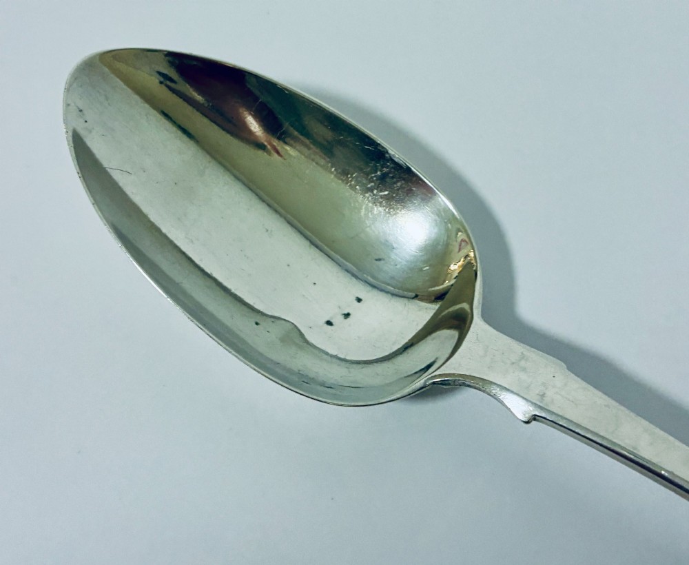 A Georgian sterling silver serving spoon featuring a polished oval bowl and a classic fiddle pattern handle resting against a plain light background. View VI