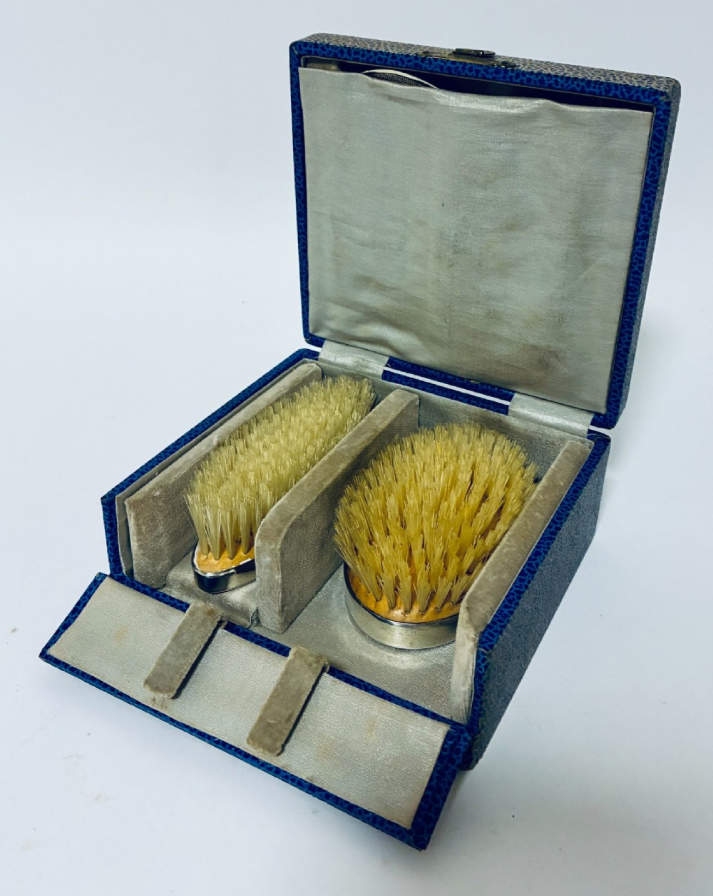 An open blue fitted presentation case displaying two silver-mounted brushes with light-coloured natural bristles against a grey velvet interior.