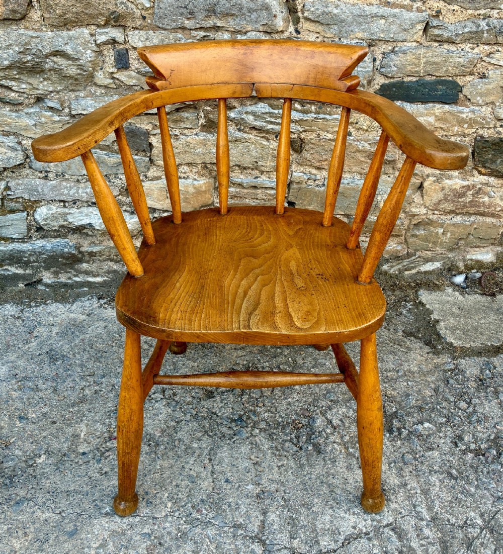 An antique elm captains armchair featuring turned spindles, a low curved backrest, a carved saddle seat, and turned legs with stretchers.