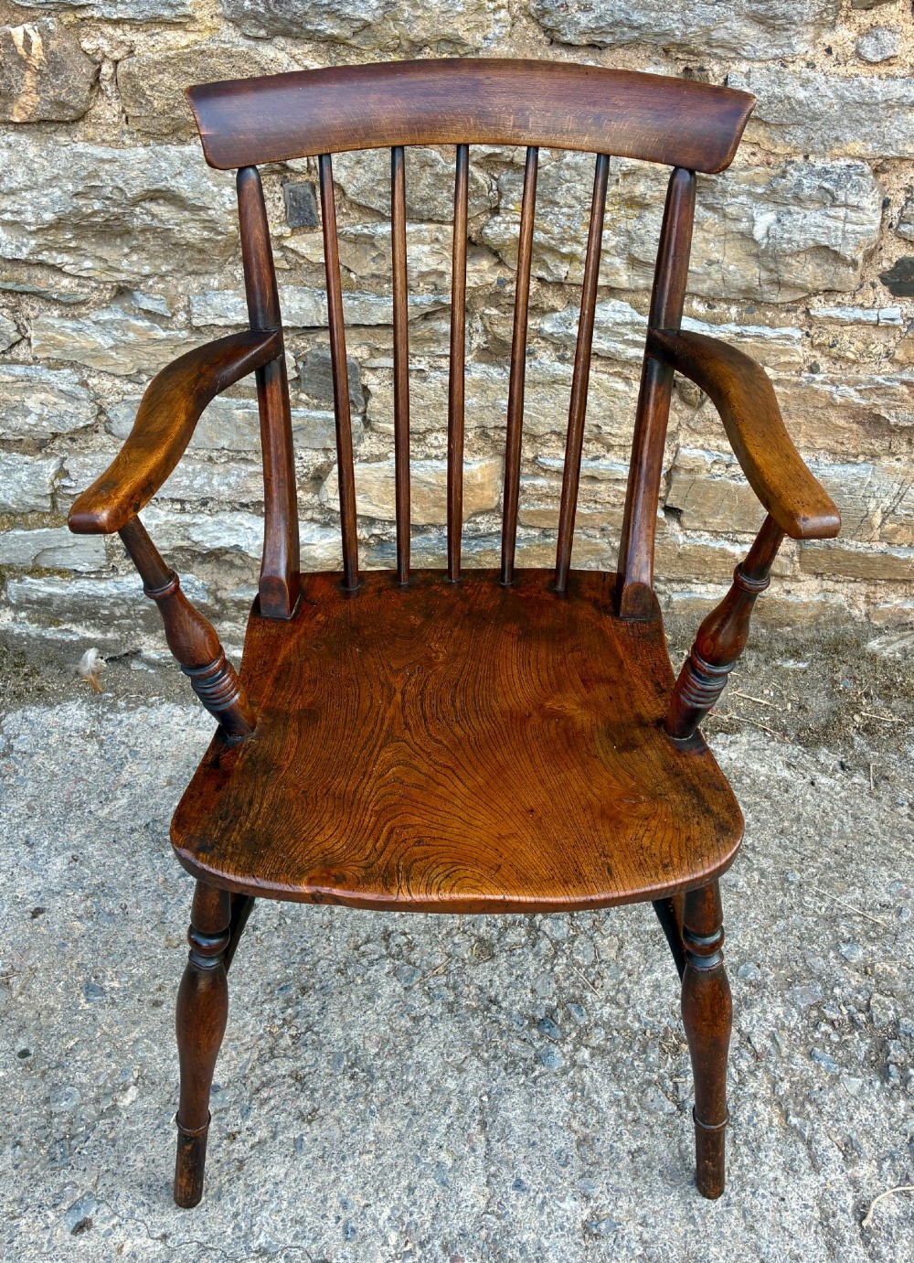 An antique dark brown wooden stick back Windsor armchair with turned legs, curved armrests, and a shaped solid elm seat.