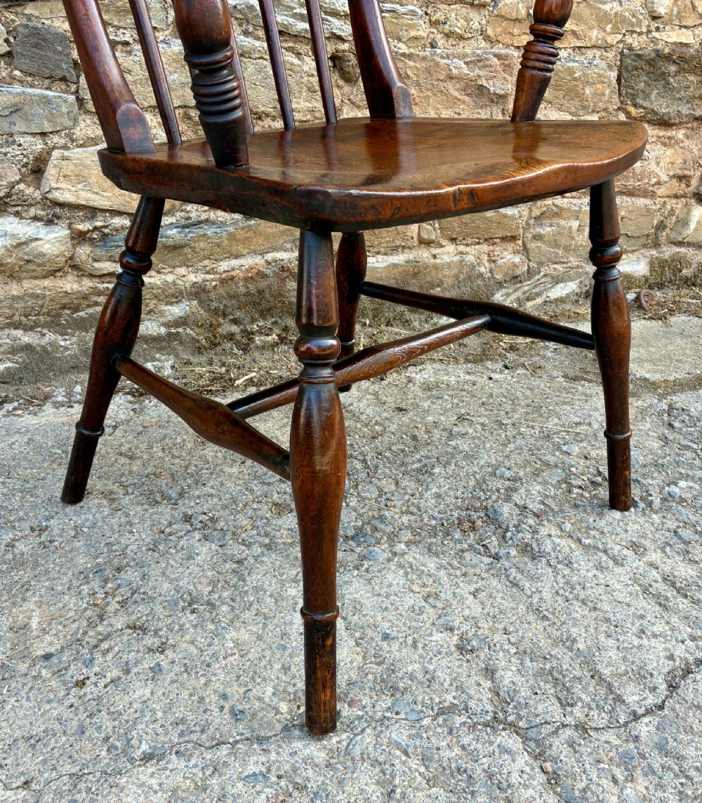 An antique dark brown wooden stick back Windsor armchair with turned legs, curved armrests, and a shaped solid elm seat. View X
