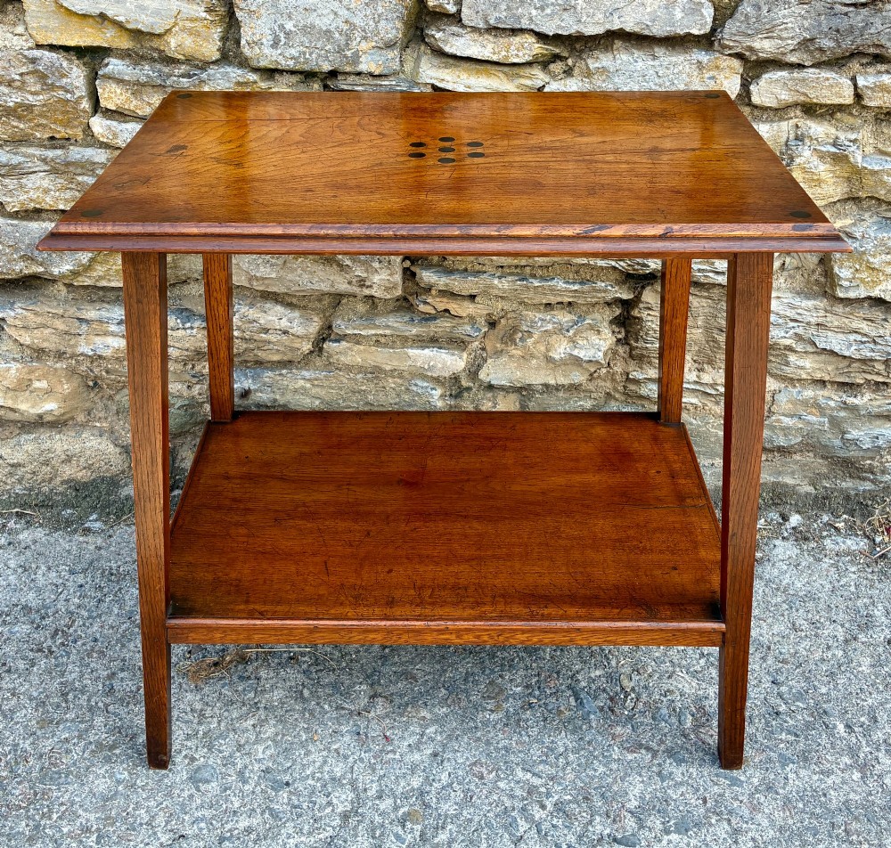 An Arts and Crafts solid oak side table featuring a lower shelf, straight square legs, and dark circular wood inlays arranged in a grid pattern on the top surface.