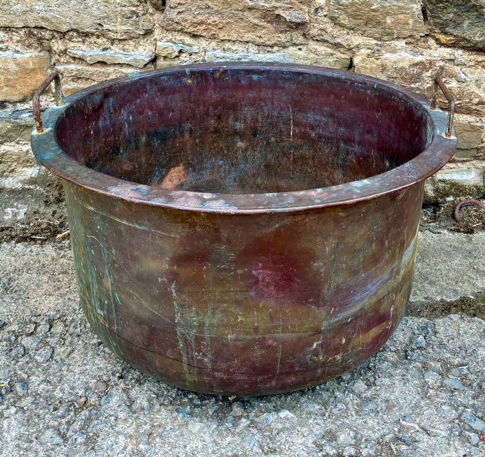 A large, circular antique copper wash tub featuring twin wrought iron carrying handles, a rolled rim, and heavy surface oxidisation with verdigris.