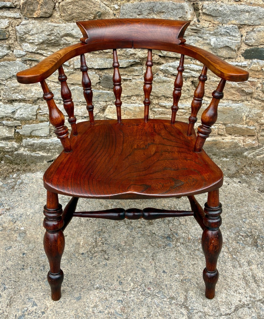 An antique warm brown elm captains chair with turned spindles, a contoured saddle seat, and turned legs connected by stretchers.