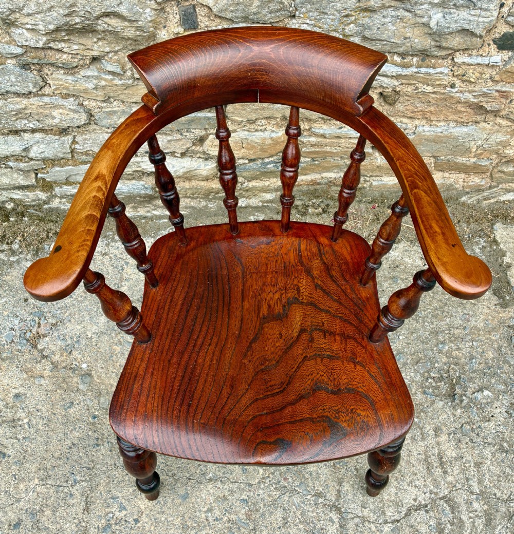 An antique warm brown elm captains chair with turned spindles, a contoured saddle seat, and turned legs connected by stretchers. View II
