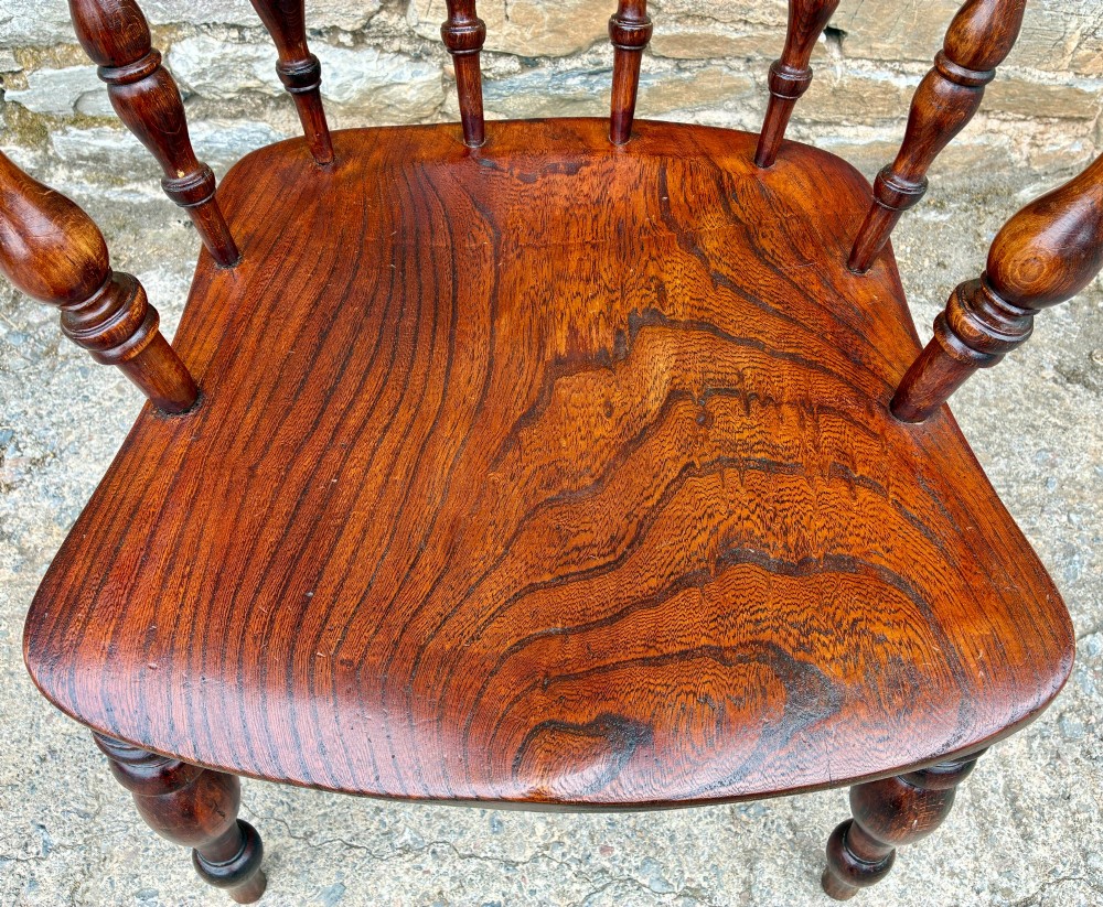 An antique warm brown elm captains chair with turned spindles, a contoured saddle seat, and turned legs connected by stretchers. View III