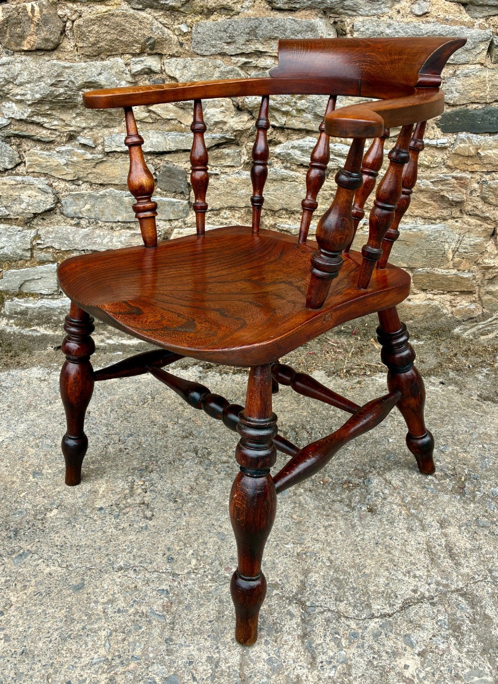 An antique warm brown elm captains chair with turned spindles, a contoured saddle seat, and turned legs connected by stretchers. View IV