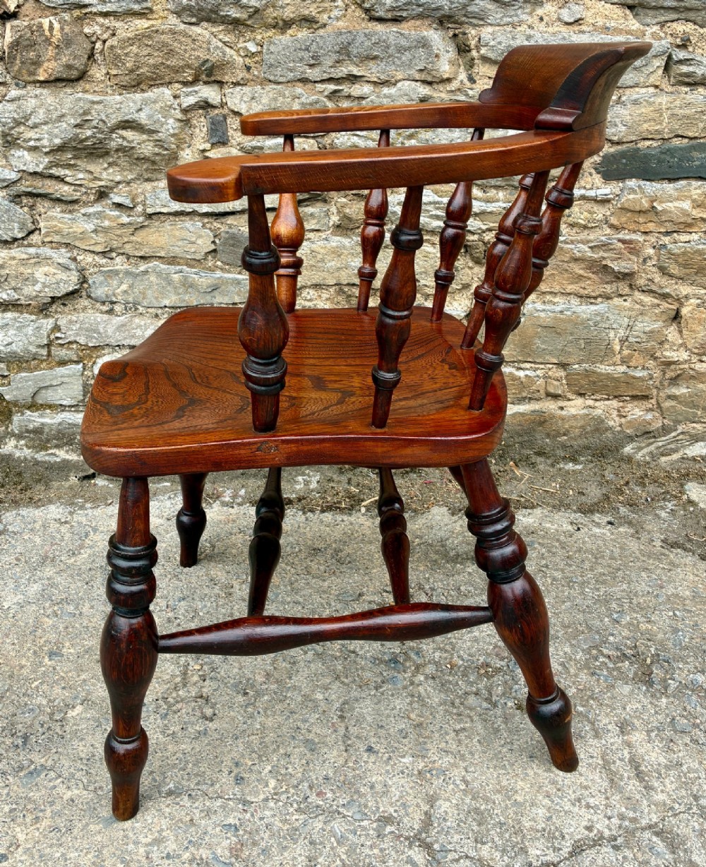 An antique warm brown elm captains chair with turned spindles, a contoured saddle seat, and turned legs connected by stretchers. View V
