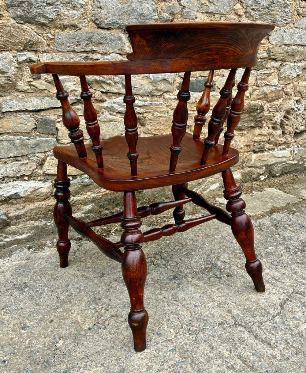 An antique warm brown elm captains chair with turned spindles, a contoured saddle seat, and turned legs connected by stretchers. View VII