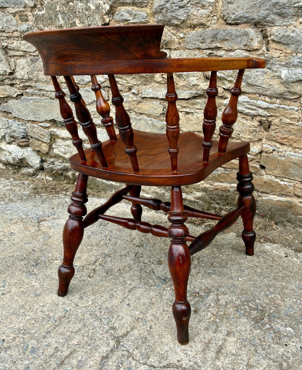 An antique warm brown elm captains chair with turned spindles, a contoured saddle seat, and turned legs connected by stretchers. View VIII