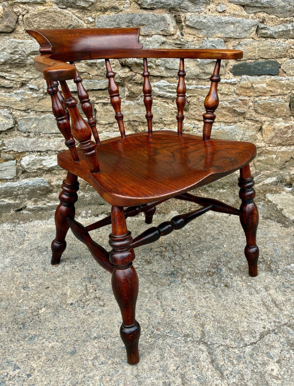 An antique warm brown elm captains chair with turned spindles, a contoured saddle seat, and turned legs connected by stretchers. View X