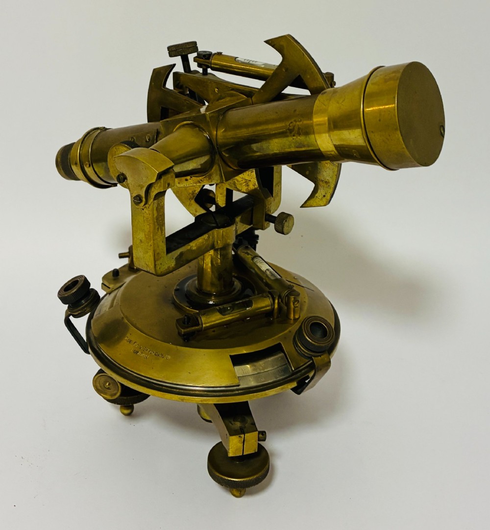 An antique brass surveyors theodolite featuring a central sighting telescope, spirit levels, and knurled adjustment screws on a circular base.