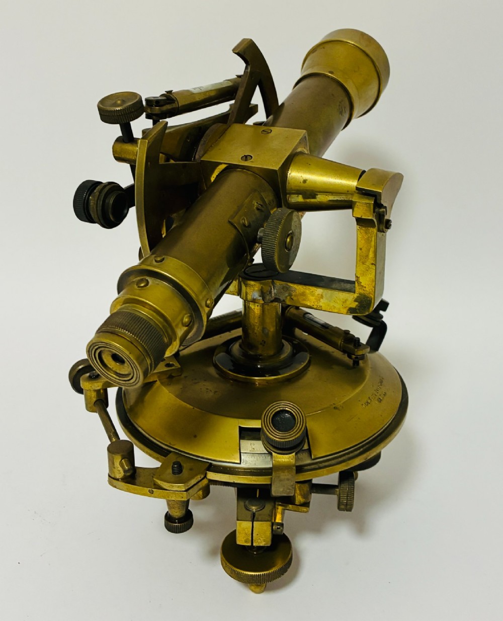 An antique brass surveyors theodolite featuring a central sighting telescope, spirit levels, and knurled adjustment screws on a circular base. View V