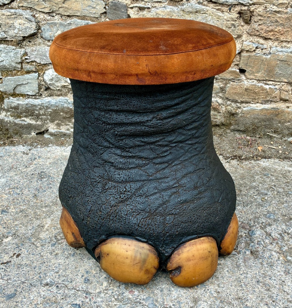 An antique taxidermy elephant foot converted into a footstool, featuring dark textured hide with preserved base toenails and a cushioned tan suede seat.