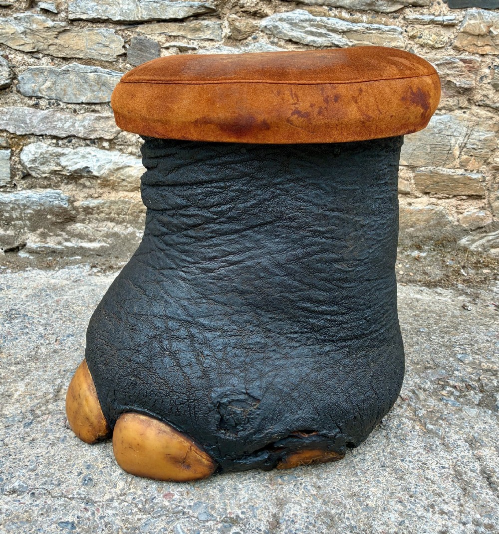 An antique taxidermy elephant foot converted into a footstool, featuring dark textured hide with preserved base toenails and a cushioned tan suede seat. View IV