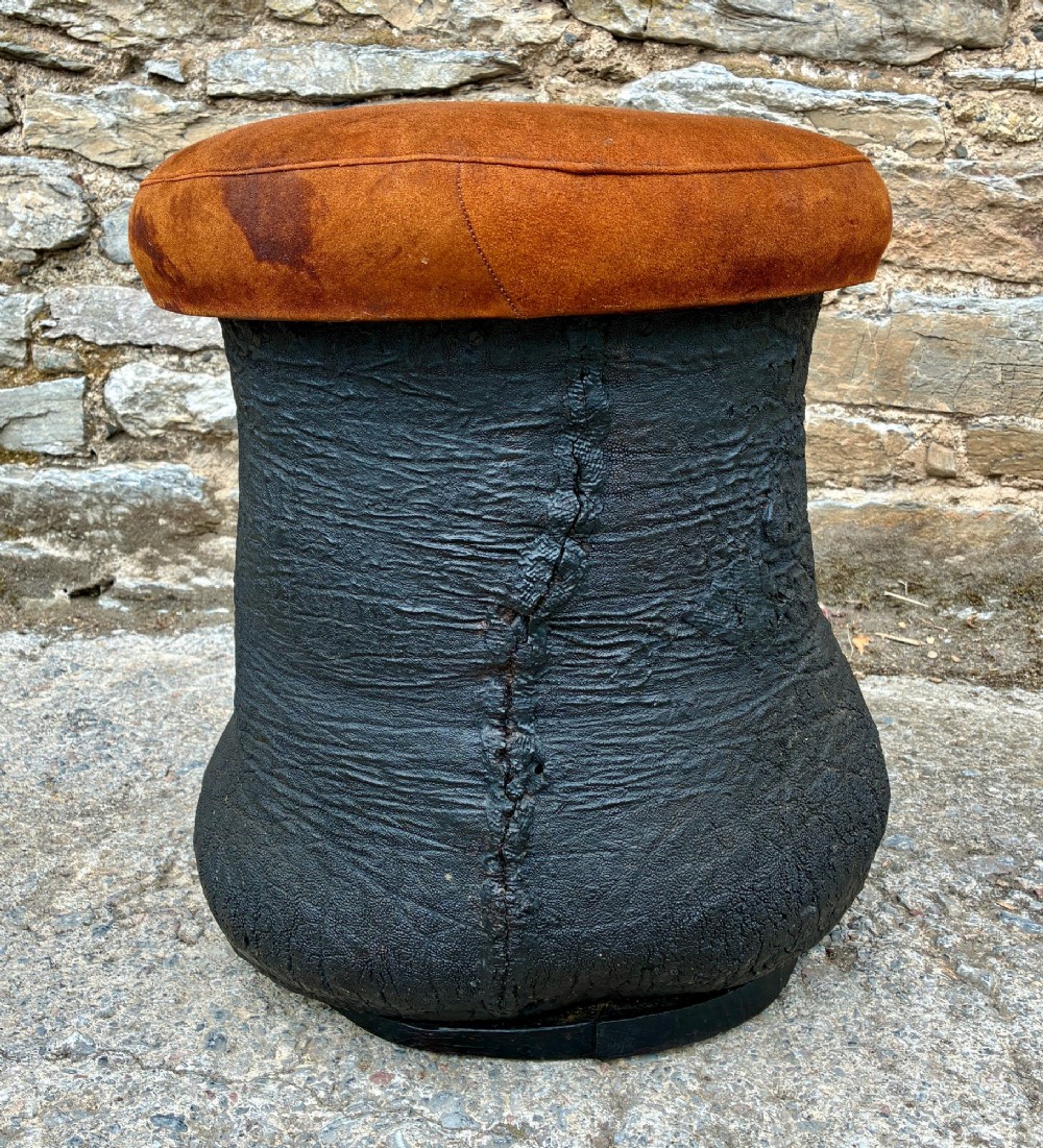 An antique taxidermy elephant foot converted into a footstool, featuring dark textured hide with preserved base toenails and a cushioned tan suede seat. View VI
