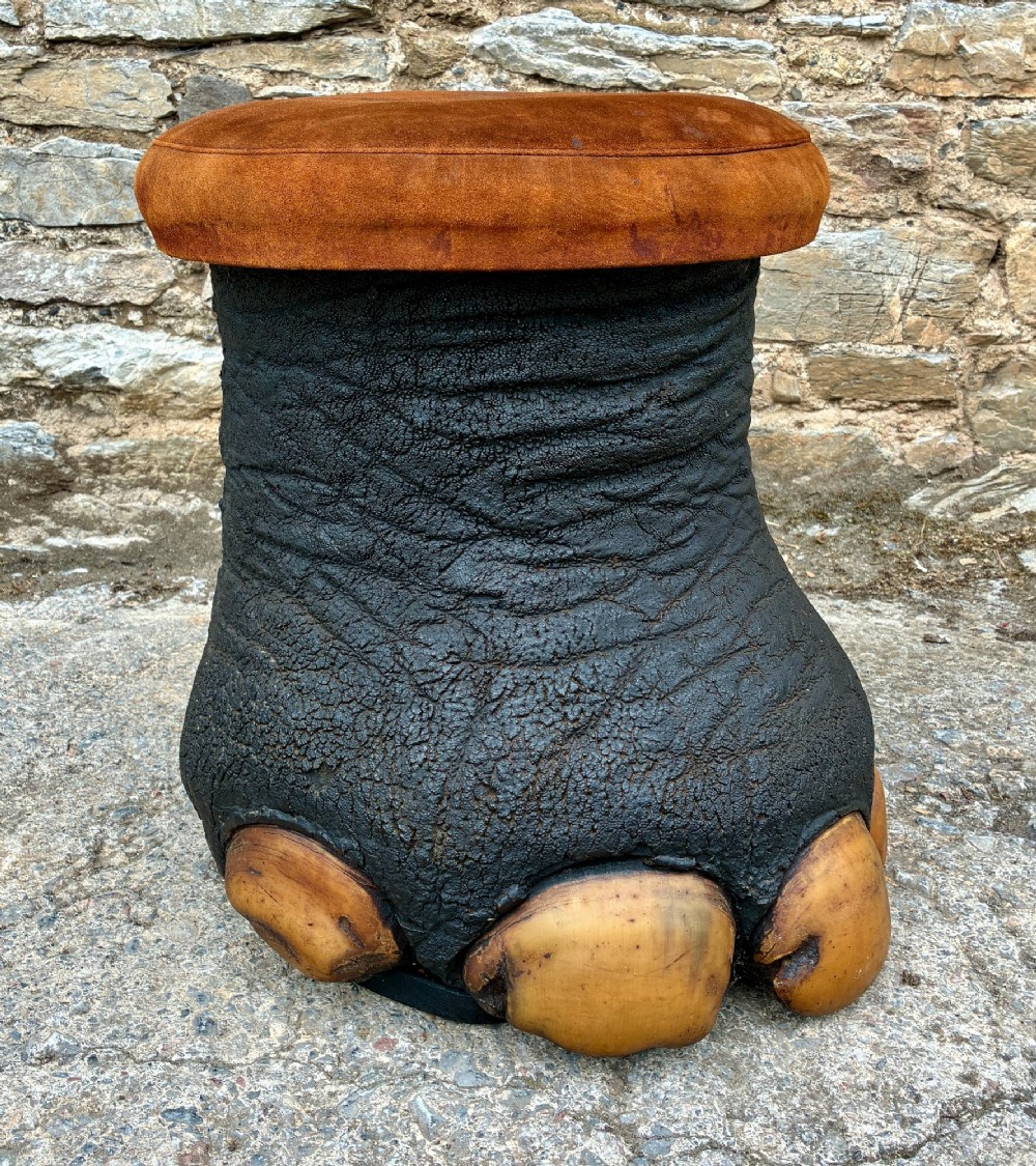 An antique taxidermy elephant foot converted into a footstool, featuring dark textured hide with preserved base toenails and a cushioned tan suede seat. View VIII