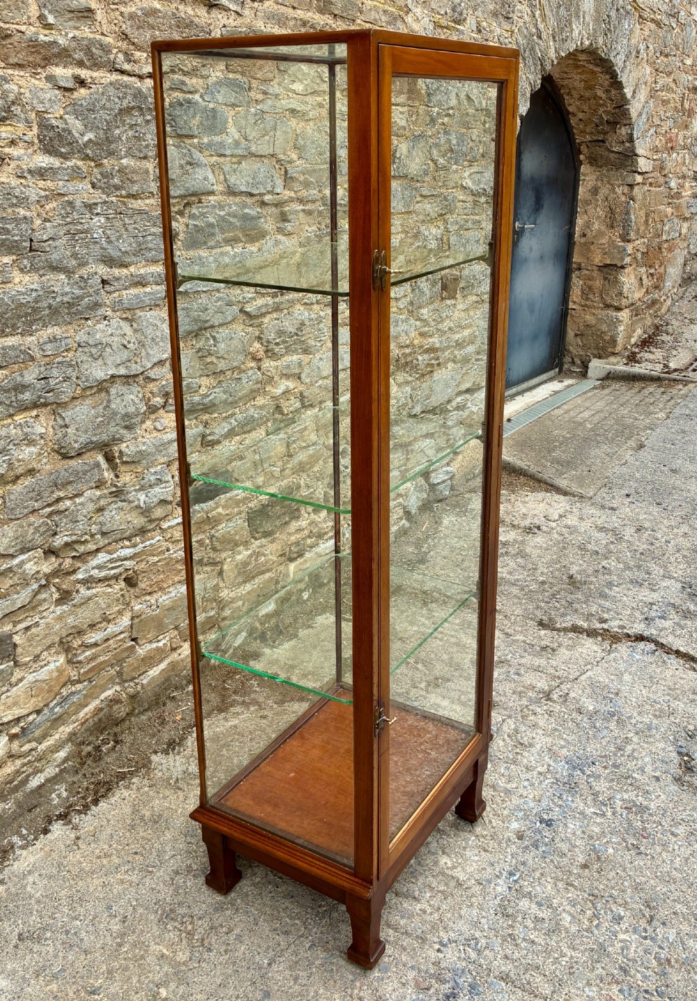 A tall, slim mahogany shop display cabinet featuring glass side panels, a single glazed door, three interior glass shelves, and brass catches. View III
