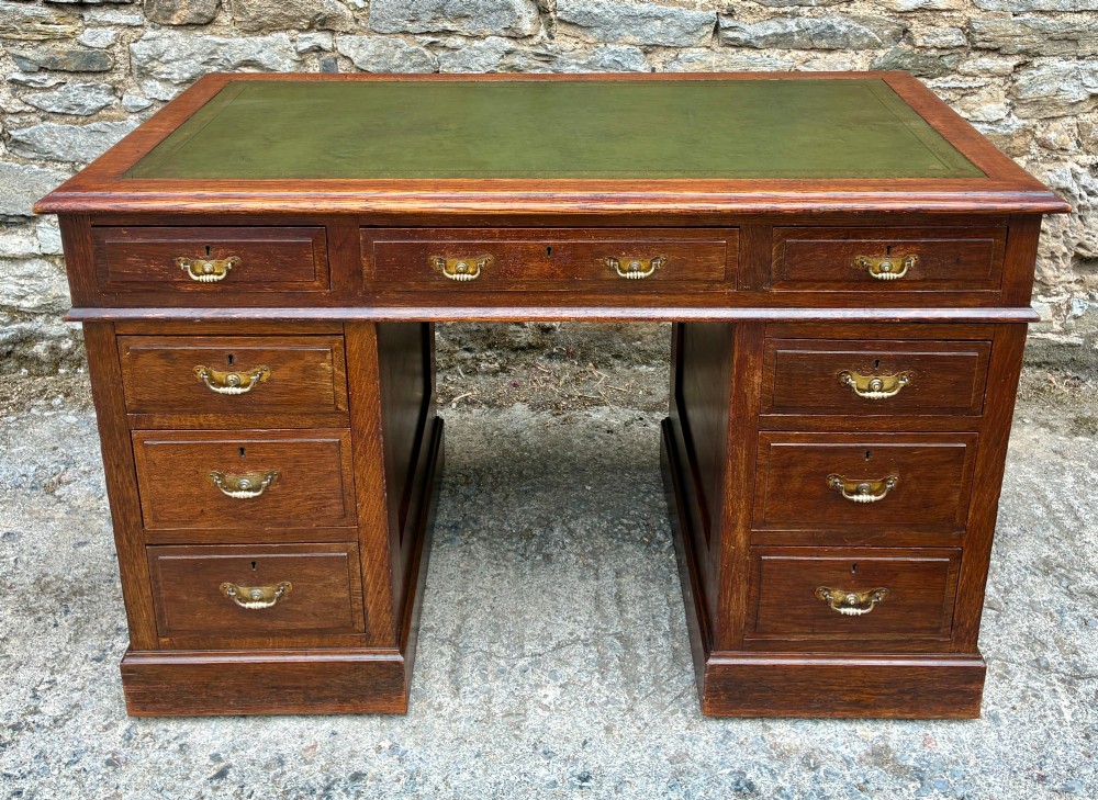 An Edwardian oak pedestal desk featuring a green leather writing surface, nine drawers with brass handles, and twin pedestals on plinth bases.