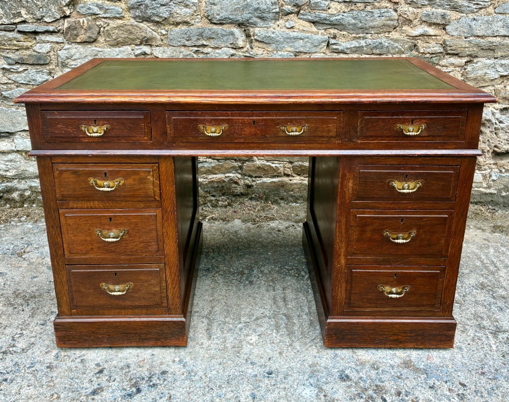 An Edwardian oak pedestal desk featuring a green leather writing surface, nine drawers with brass handles, and twin pedestals on plinth bases. View II